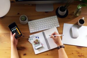 A person's hand holding a phone with a calculator display, while writing notes next to cash, a coffee cup, and a computer keyboard.