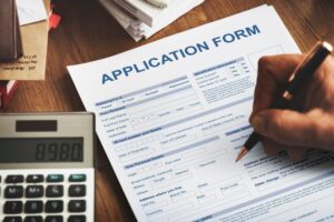 A hand holds a pen, filling out an application form on a wooden desk with a calculator and scattered papers.
