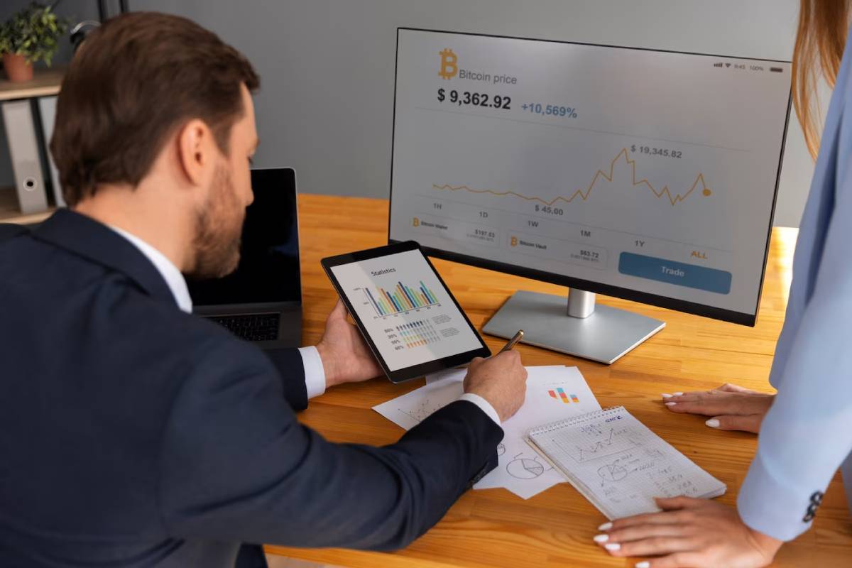 A businessman analyzes Bitcoin price data on a tablet, while a chart displays trends on a monitor, with notes scattered on the table.
