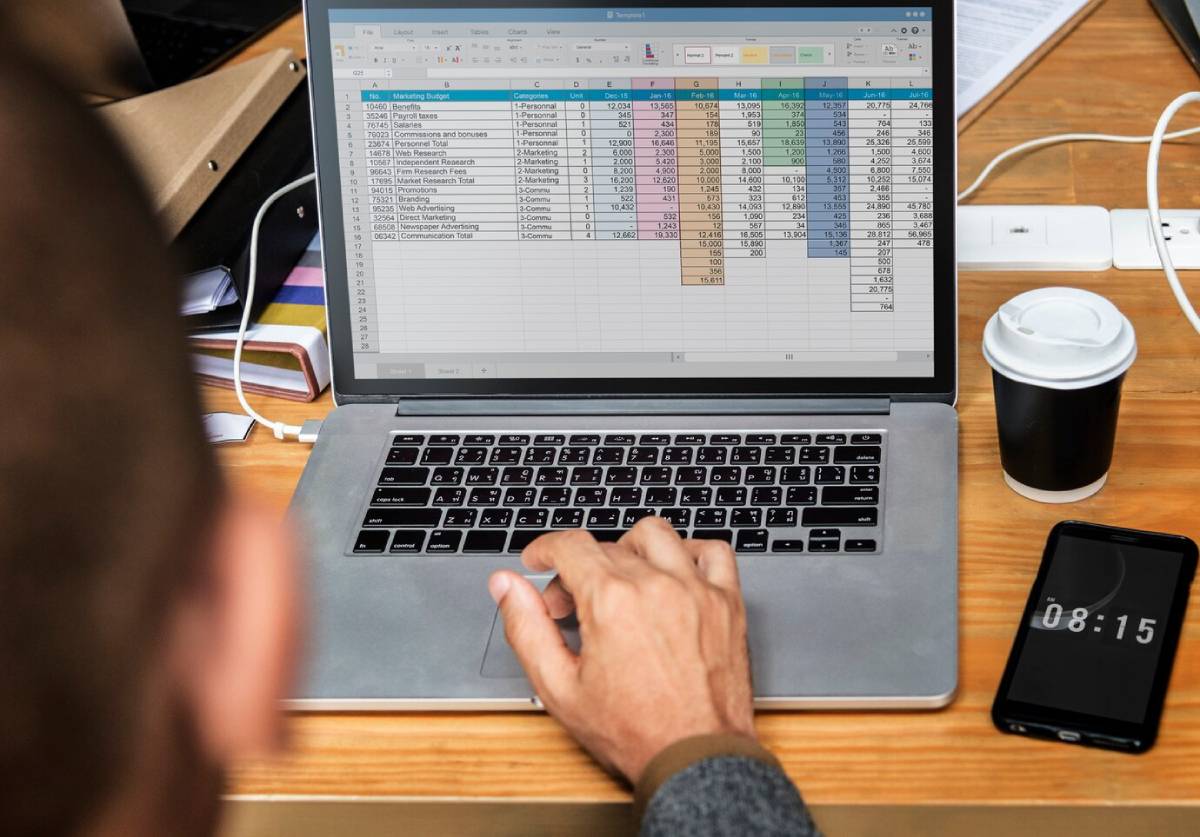 A person uses a laptop to work on a marketing budget spreadsheet, with a coffee cup and smartphone displaying the time nearby. 