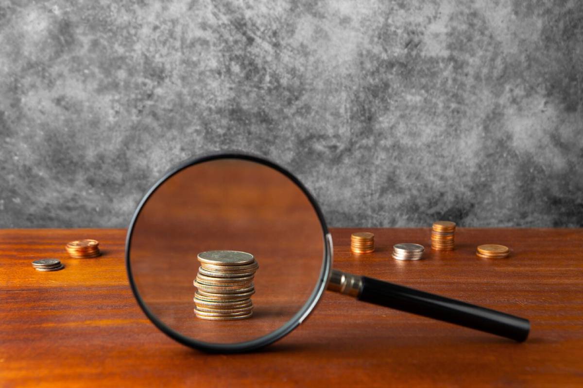 A magnifying glass focuses on a stack of coins, highlighting them against a background of scattered coins and a textured gray surface.