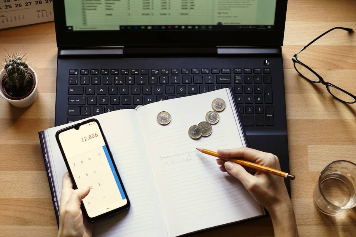 A person uses a calculator on their phone while writing notes in a notebook beside coins and a glass of water, with a laptop in view. 