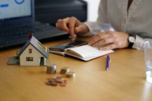 A person's hand using a calculator beside a model house, coins, a notebook, and a pen on a wooden desk with a laptop in the background.