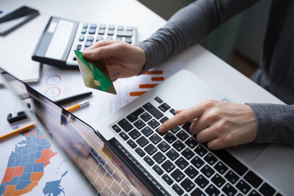 A person with a credit card in hand is using a laptop to make an online transaction, surrounded by office supplies and a calculator.