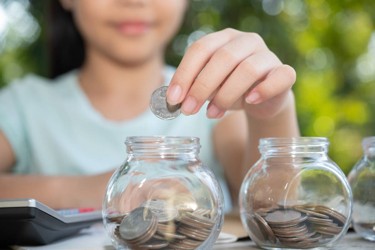 A girl deposits a coin into a glass jar filled with coins, focused on saving money, with a calculator and greenery in the background. 