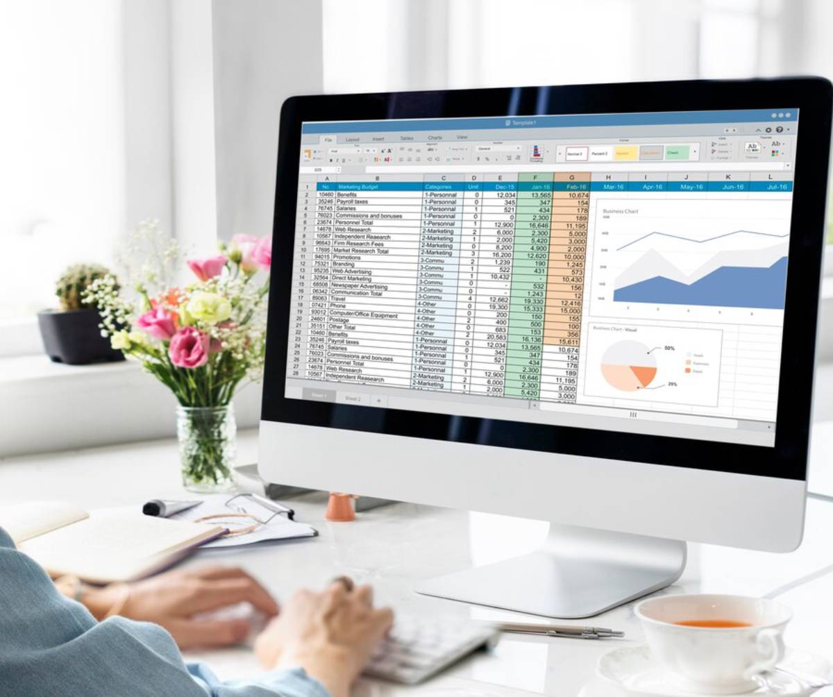 A person works on a spreadsheet displaying a marketing budget, with colorful charts and a vase of flowers on the desk. 