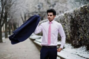 A person wearing a pink shirt and a pink tie confidently tosses a dark blue suit jacket, set against a wintery landscape.