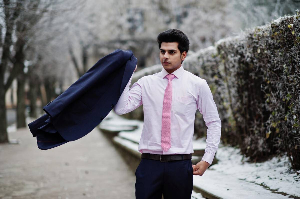 A person wearing a pink shirt and a pink tie confidently tosses a dark blue suit jacket, set against a wintery landscape.