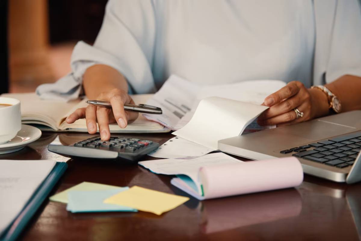 A person calculates expenses using a calculator, surrounded by paperwork, a laptop, sticky notes, and a cup of coffee.