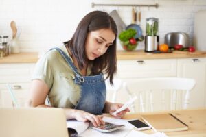 A person in a cozy kitchen calculates expenses with a calculator, surrounded by notebooks, a smartphone, and fresh produce.