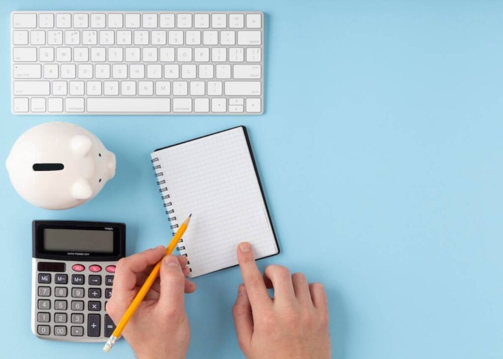 Hands holding a pencil over a blank notebook, with a piggy bank and calculator nearby, set on a light blue background.