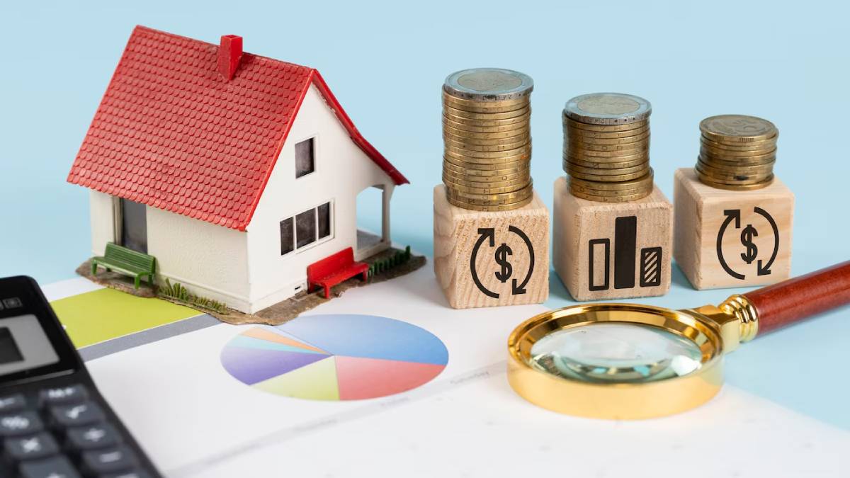  A miniature house sits next to stacks of coins and wooden blocks with financial symbols, alongside a calculator and a magnifying glass.