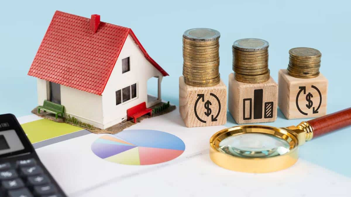 A miniature house sits next to stacks of coins and wooden blocks with financial symbols, alongside a calculator and a magnifying glass.