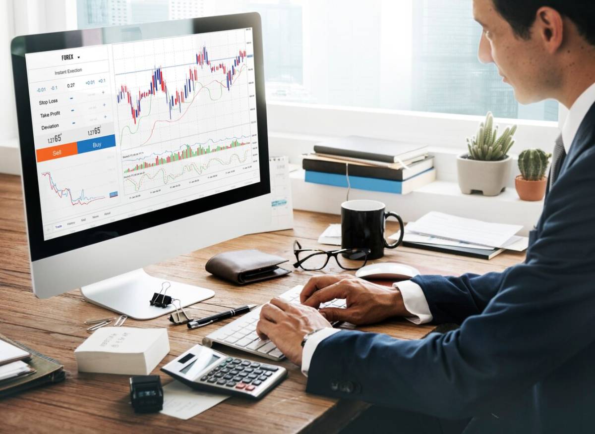 A person in a suit analyzes financial charts on a computer screen, surrounded by office supplies and a coffee mug.