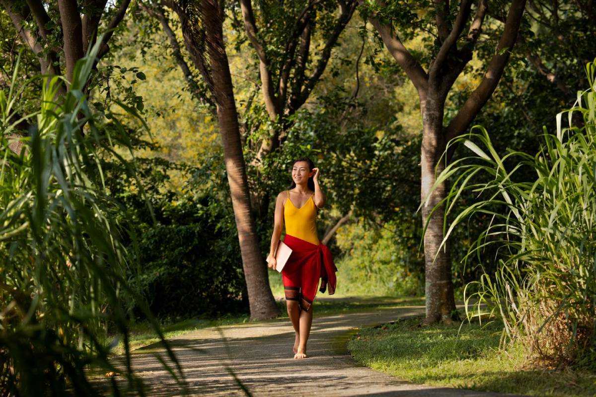A person in a yellow top and red wrap walks along a winding path surrounded by lush greenery and tall trees.