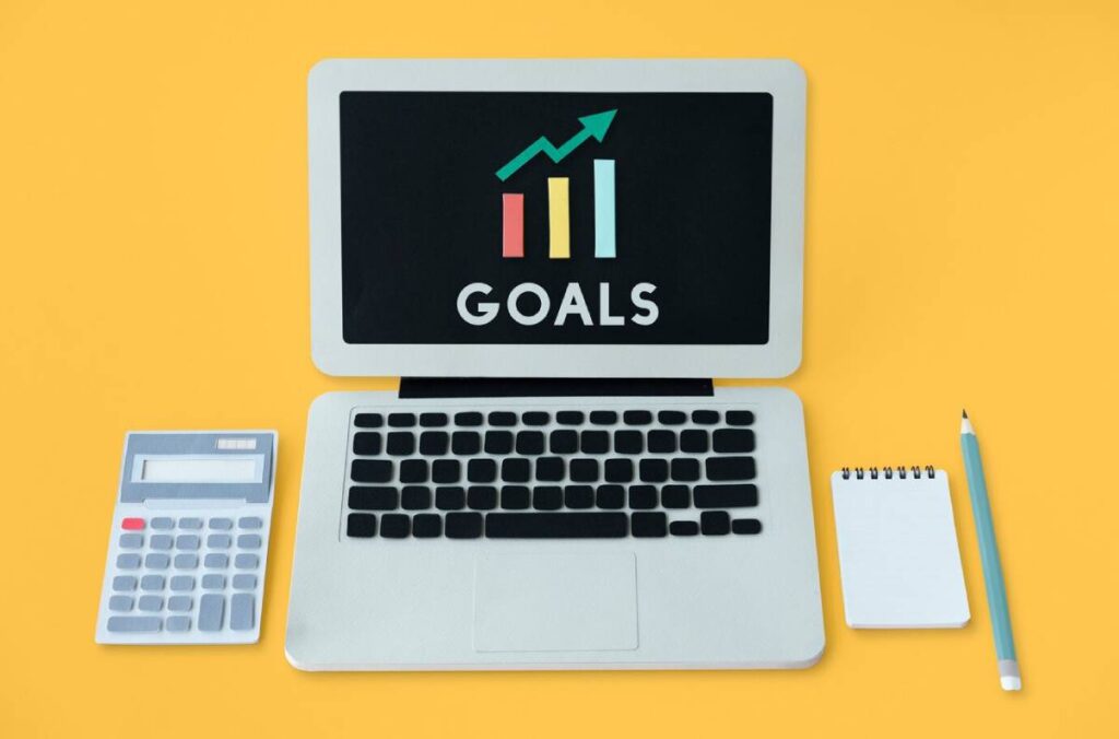 A laptop displaying GOALS with a growth chart, alongside a calculator, notepad, and pencil, set against a yellow background.