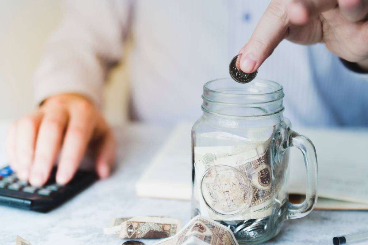 A person drops a coin into a jar filled with cash, while using a calculator, symbolizing saving or budgeting.