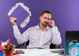 A man in a white shirt holds a purple folder with cash while sitting at a desk with a laptop and blue clock, against a purple background.