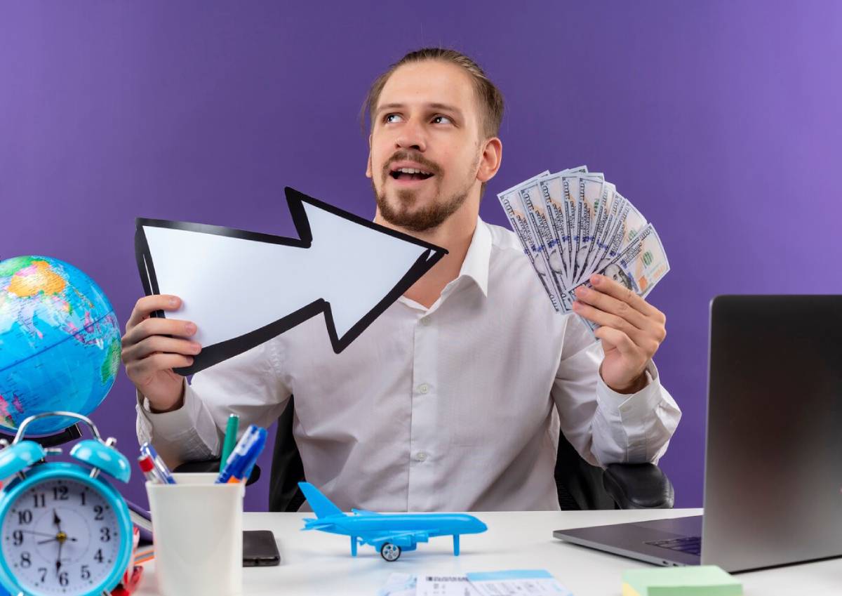 A businessman holds cash and an arrow sign while seated at a desk with a globe, clock, and model airplane, against a purple backdrop. 