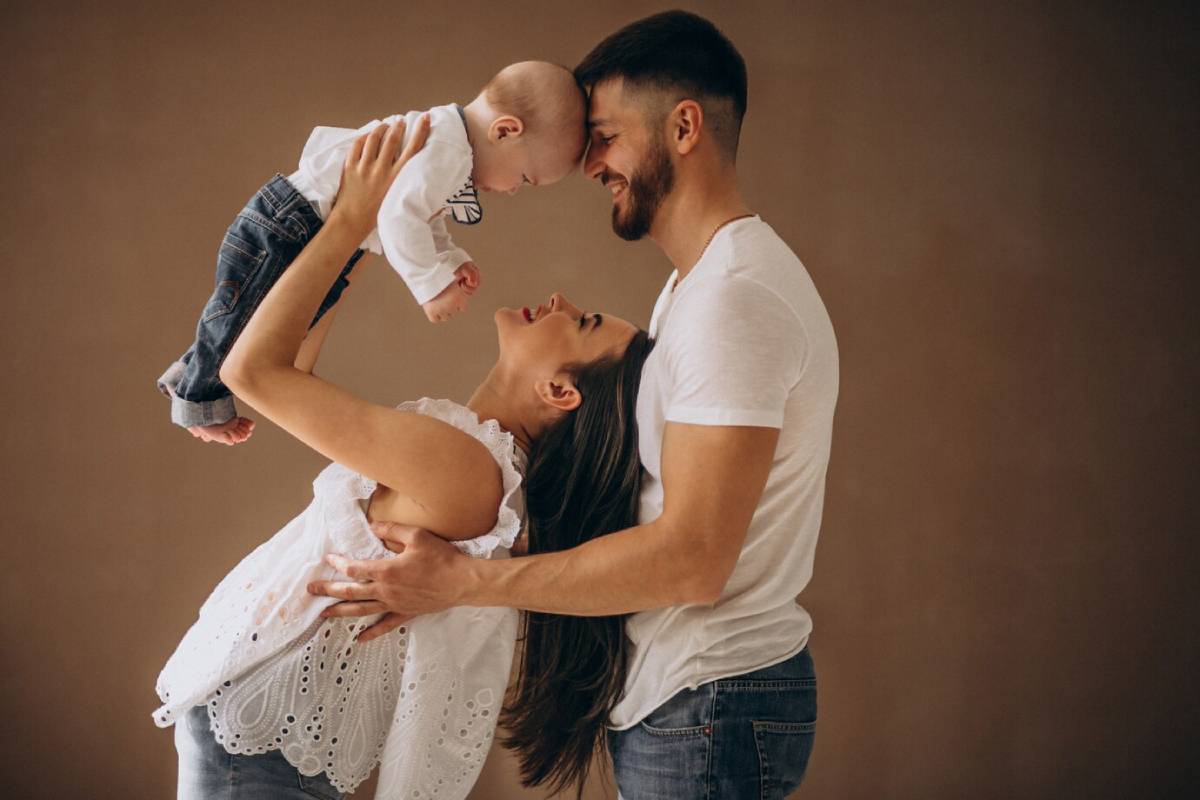 A joyful mother lifts her baby in the air, while a father watches lovingly, all set against a warm, neutral background.