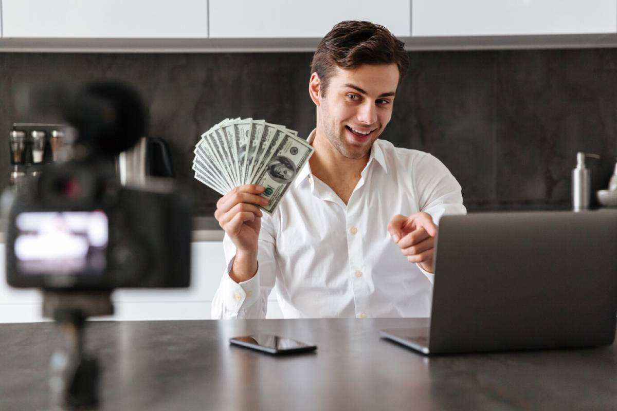 A man in a white shirt holds stacks of cash while pointing towards a laptop, with a camera capturing his engagement. 