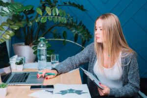 A person with long hair sits at a desk with a laptop, holding a clipboard, surrounded by plants and bottles of water.