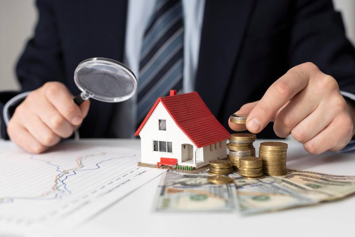 A person in a suit uses a magnifying glass on a miniature house surrounded by stacks of coins and bills, symbolizing real estate investment.