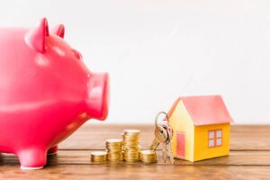A pink piggy bank sits near stacks of coins, a yellow paper house, and keys, symbolizing savings for home ownership.