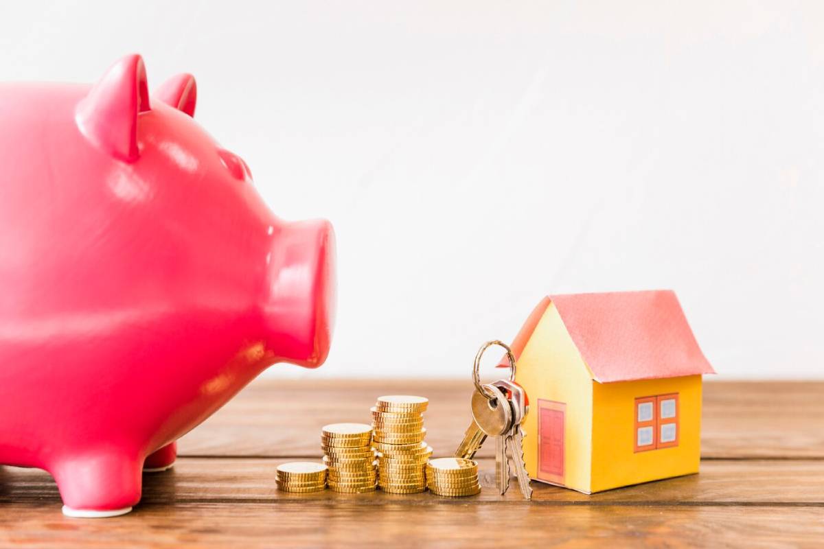A pink piggy bank sits near stacks of coins, a yellow paper house, and keys, symbolizing savings for home ownership.