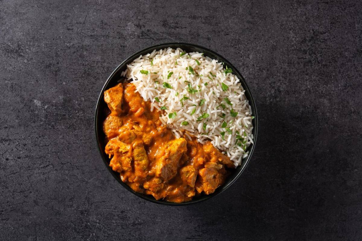  Bowl of creamy chicken curry next to fluffy white rice, garnished with fresh herbs on a dark textured background.