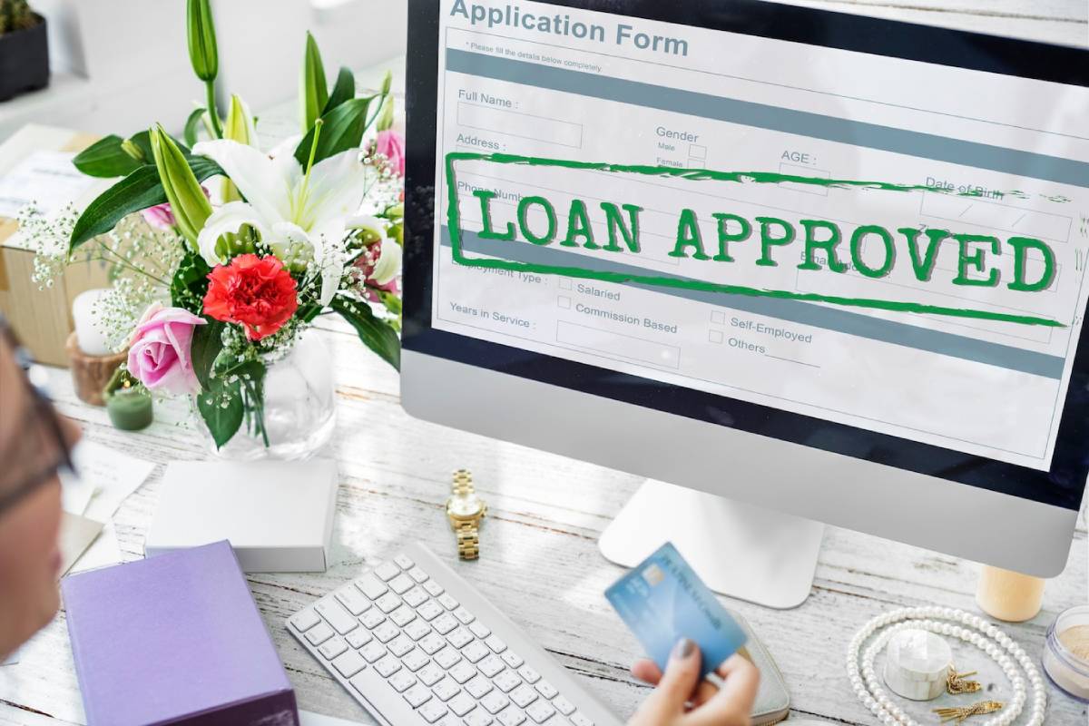 A person holds a credit card in front of a computer displaying an approved loan application, with a bouquet of flowers nearby. 