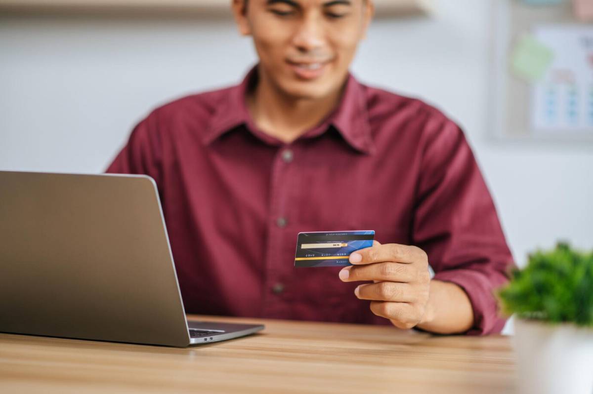 A person in a maroon shirt holds a credit card, sitting in front of a laptop on a wooden table, surrounded by greenery.