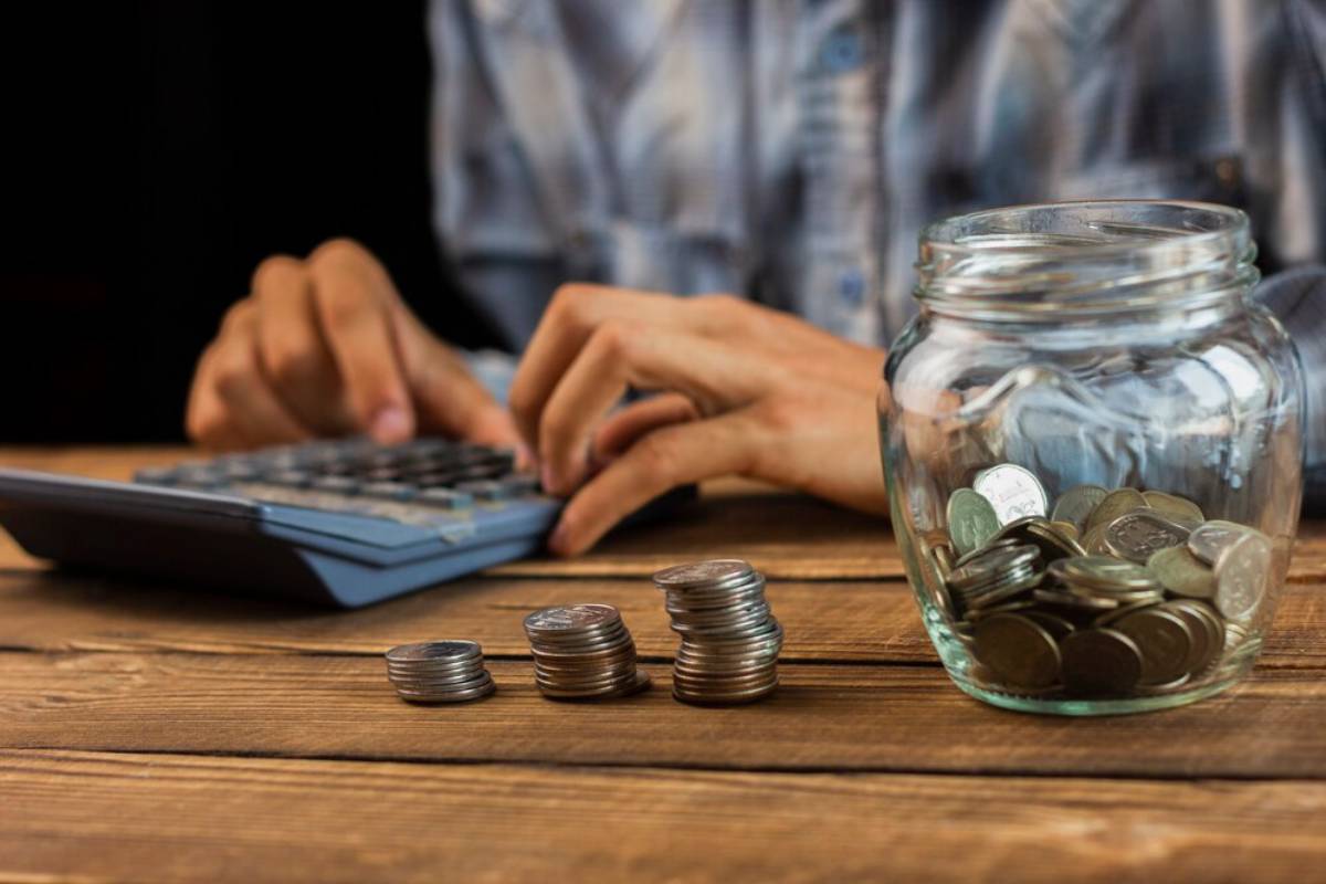 A person using a calculator next to stacked coins and a jar filled with coins on a wooden table, focusing on budgeting or finances.