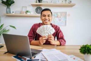 A person in a burgundy shirt holds a fan of dollar bills in front of a laptop on a desk filled with papers and a small plant.