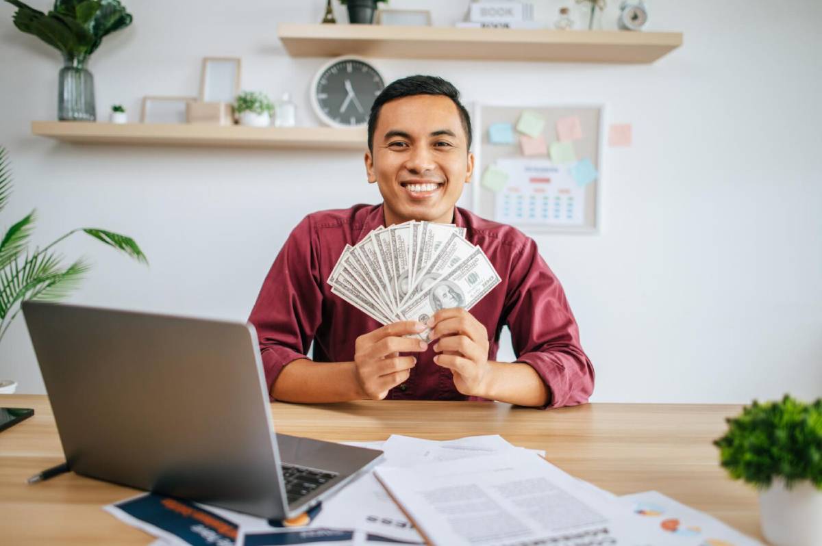 A person in a burgundy shirt holds a fan of dollar bills in front of a laptop on a desk filled with papers and a small plant.