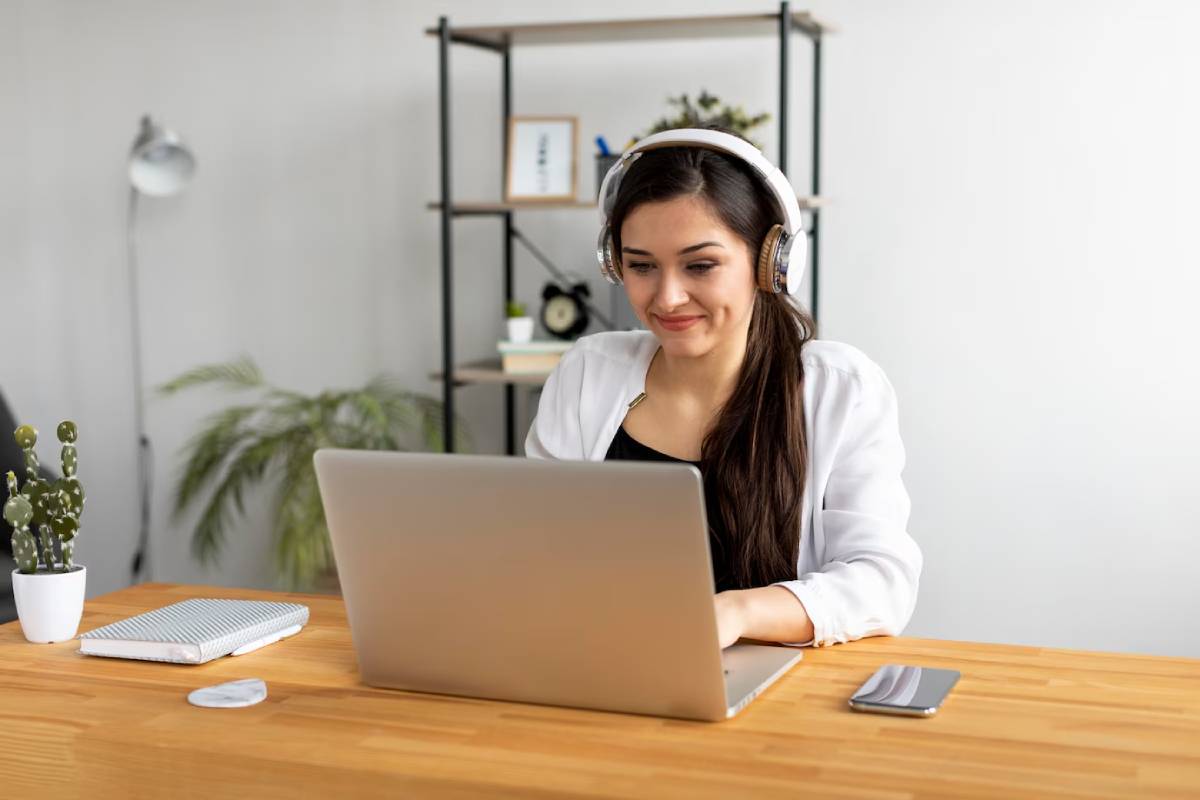A person wearing headphones sits at a wooden desk, focused on a laptop, with plants and a notebook in a modern workspace. 
