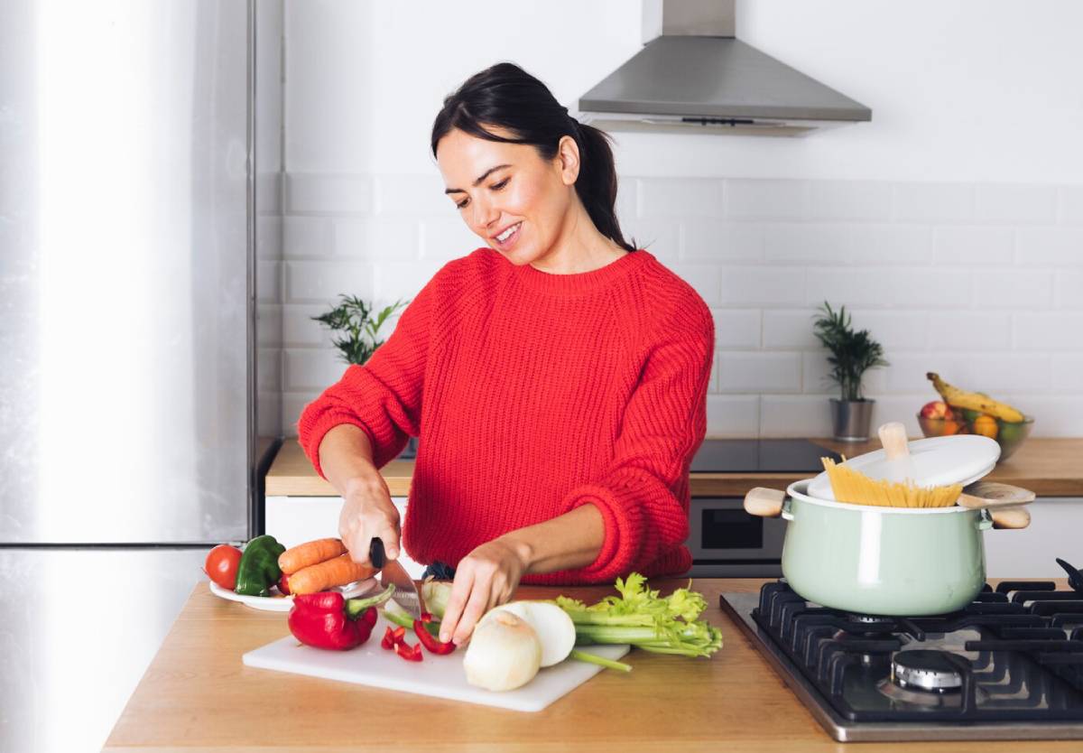 A person in a red sweater chops vegetables on a cutting board in a modern kitchen, with pasta boiling nearby.