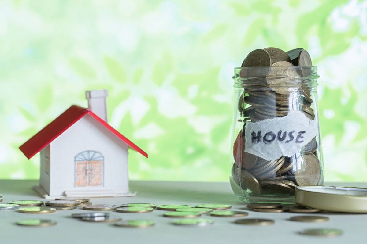 A small paper house next to a jar labeled HOUSE filled with coins on a blurred green background, symbolizing savings for a home.
