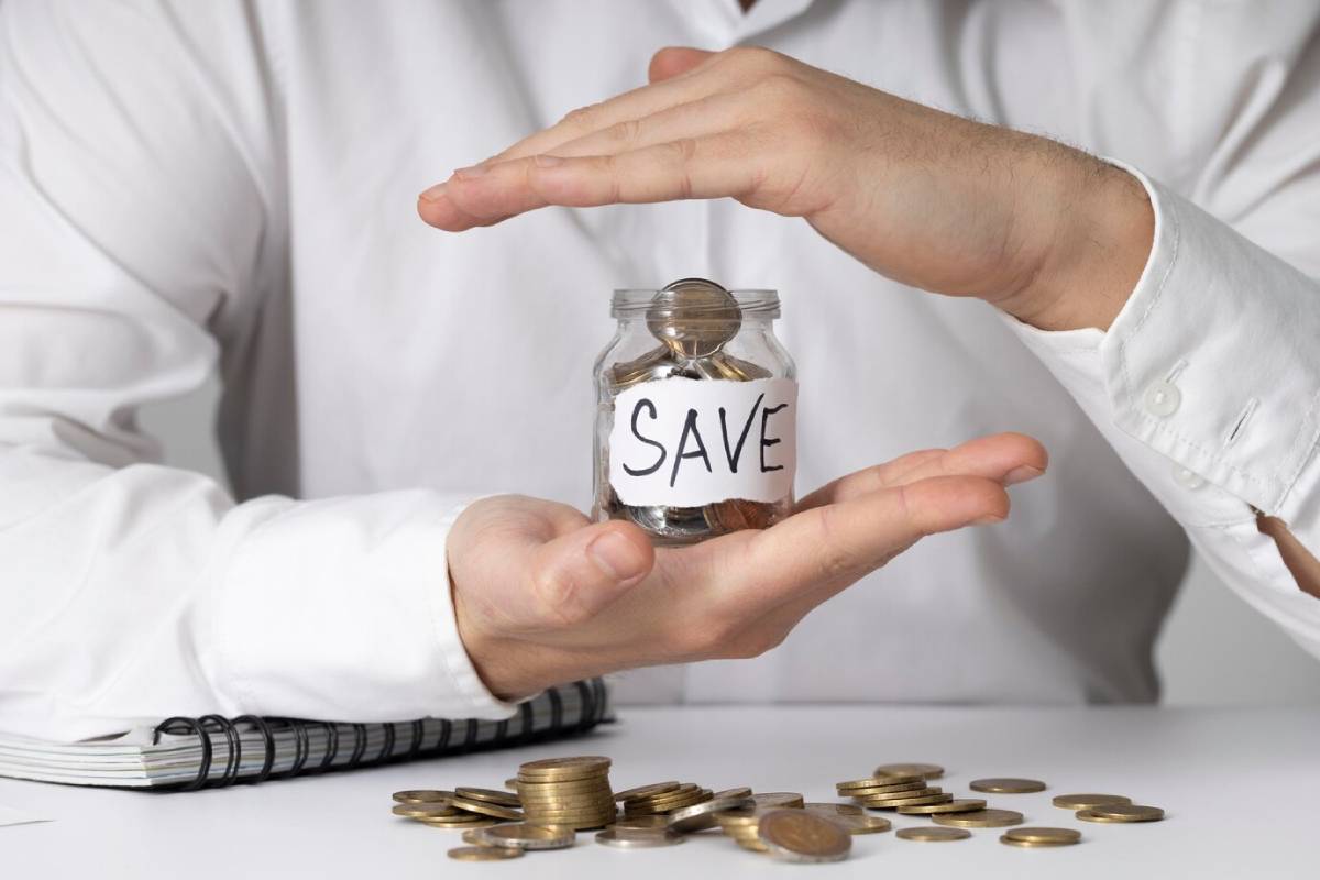 A person in a white shirt holds a jar labeled SAVE filled with coins, with more coins scattered on the table.