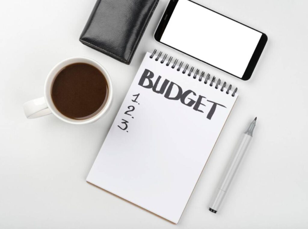 A notepad labeled BUDGET with numbered lists, a coffee cup, a pen, a black wallet, and a blank smartphone on a white background.