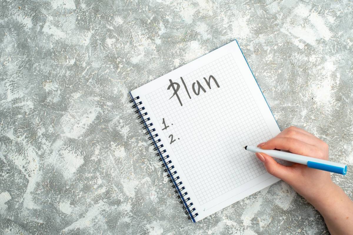 A hand holding a pen over a notebook with the word Plan written at the top and two numbered lines for notes. 