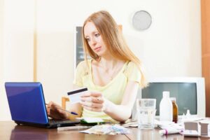 A woman in a light yellow top sits at a cluttered table, using a laptop and holding a credit card, surrounded by various items.