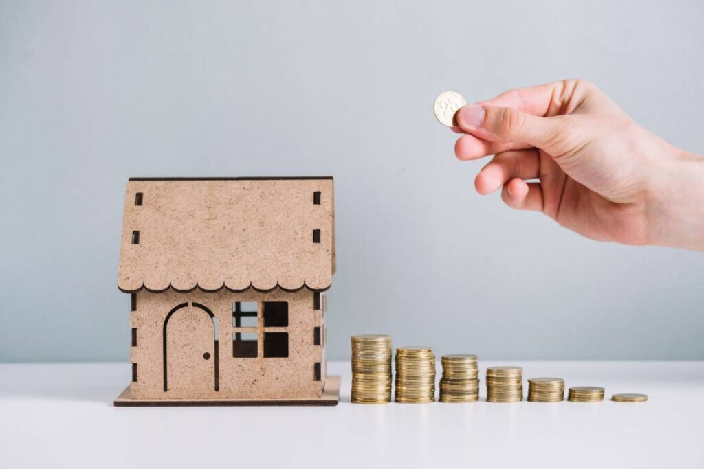 A hand is placing a coin next to a stacked arrangement of coins beside a miniature wooden house model.