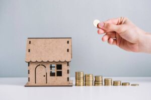 A hand is placing a coin next to a stacked arrangement of coins beside a miniature wooden house model.