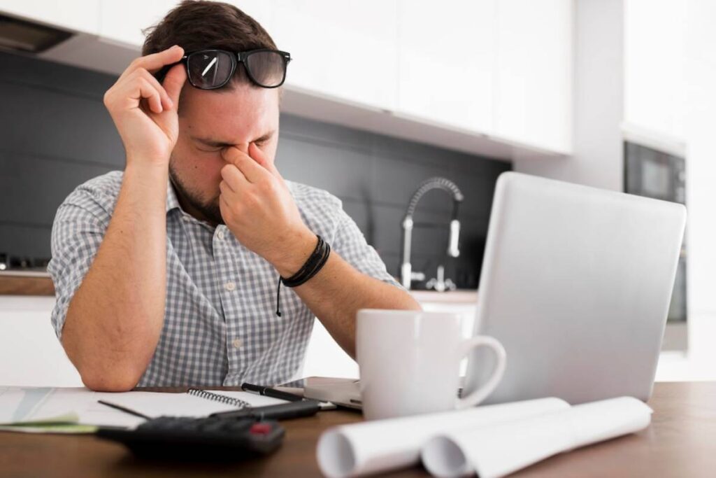 A person sits at a desk, wearing sunglasses, with a laptop open, a coffee mug, and notes scattered around in a modern kitchen.