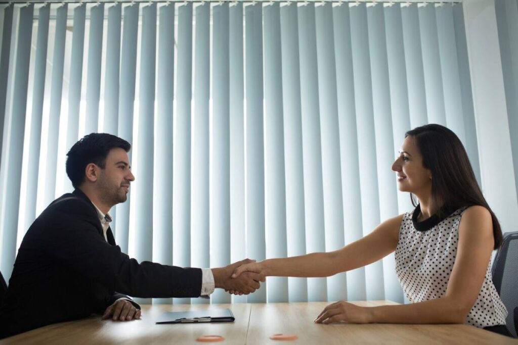 A professional handshake between two individuals at a table, with vertical blinds filtering sunlight, symbolizing agreement or partnership.