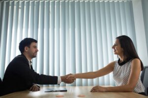 A professional handshake between two individuals at a table, with vertical blinds filtering sunlight, symbolizing agreement or partnership.