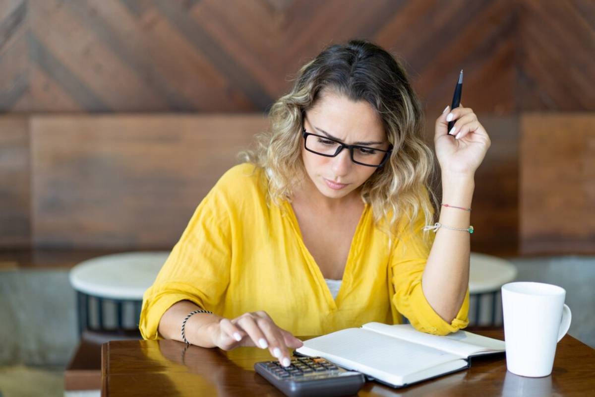 A woman in a bright yellow shirt sits at a wooden table, using a calculator with a notebook and a white mug beside her.