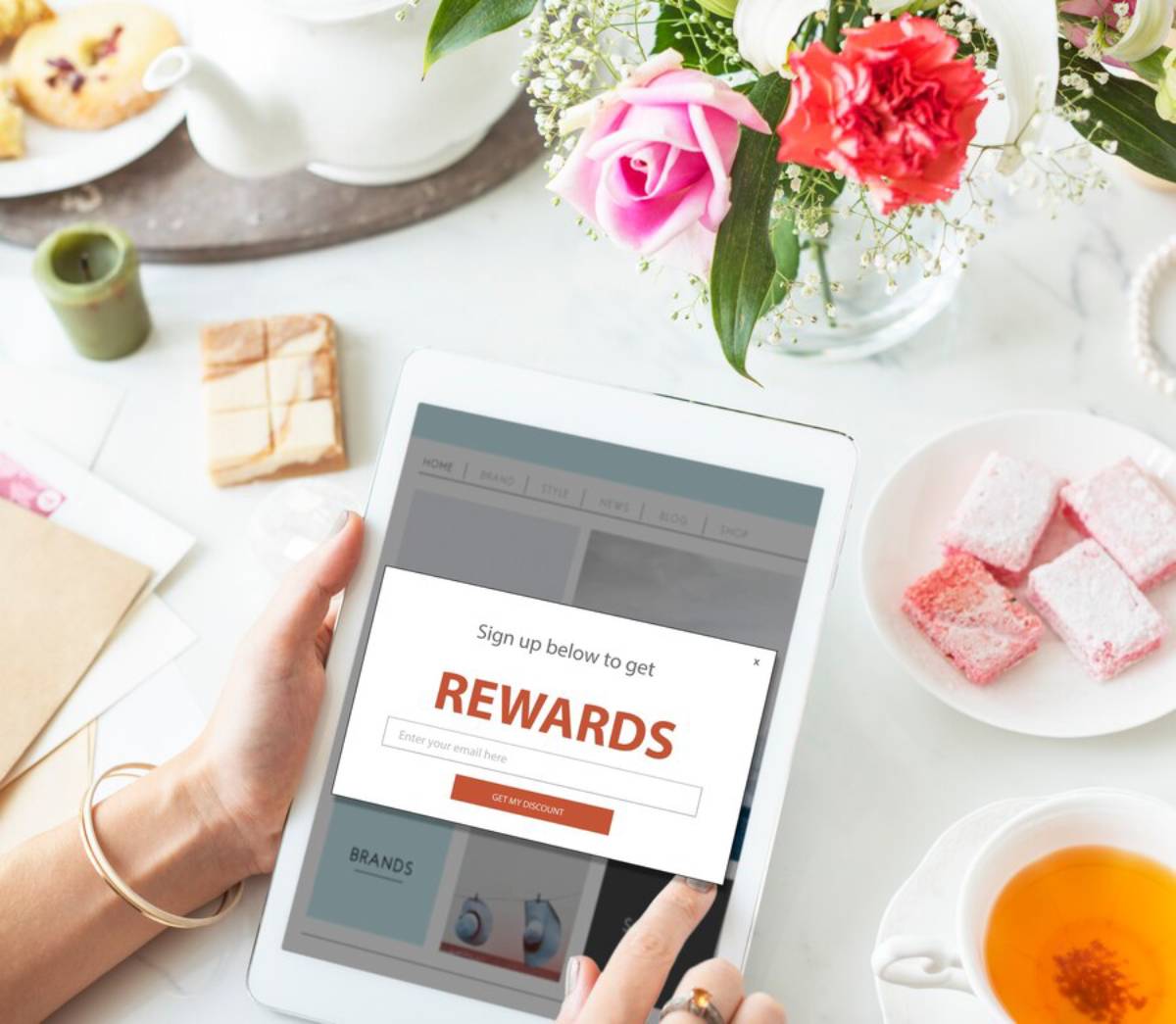 A person using a tablet to sign up for rewards, surrounded by tea, pastries, flowers, and a candle on a table.