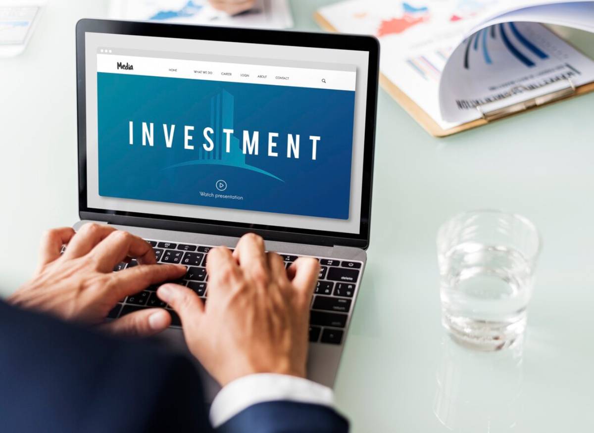 A person typing on a laptop displaying a blue INVESTMENT webpage, with a glass of water and documents on the desk. 